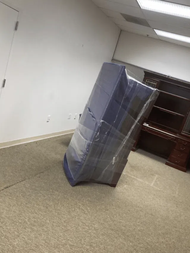 Office room with carpet, wrapped mattress leaning against wall near dark wooden desk and closed white door.