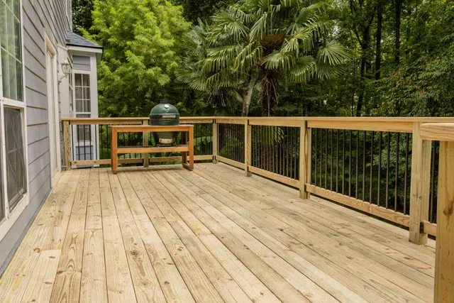 Marietta spacious wooden deck attached to house with railing and green ceramic grill on table against lush trees.