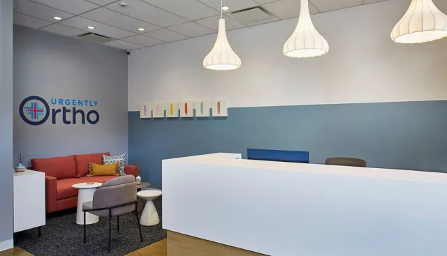 Modern orthopedic clinic reception area with white desk, hanging lights, red sofa, and wall art under bright lighting.