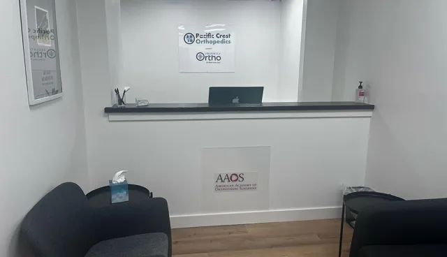 Modern orthopedic clinic reception area with gray chairs, tissue box, laptop, and medical organization logos on white walls.
