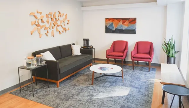 Modern living room with dark gray sofa, red chairs, geometric wall art, white round table, and a large patterned rug