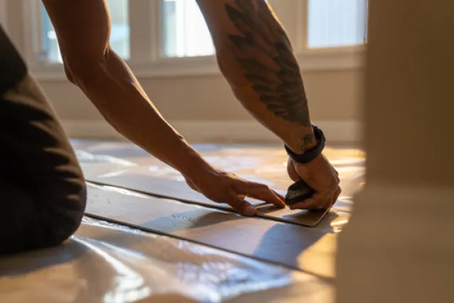 Person installing vinyl flooring with cutting tool in a sunlit room, showing arm tattoos and focused work.