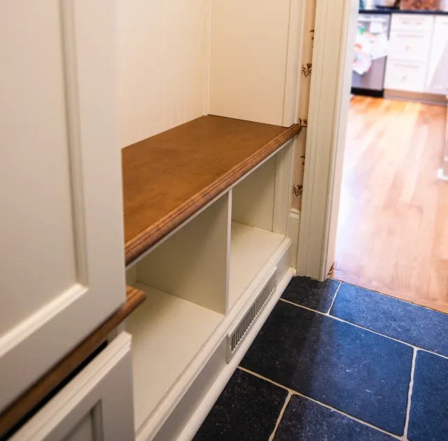 Built-in white storage bench with wooden seat and cubbies in hallway next to kitchen with tile and wood floors.