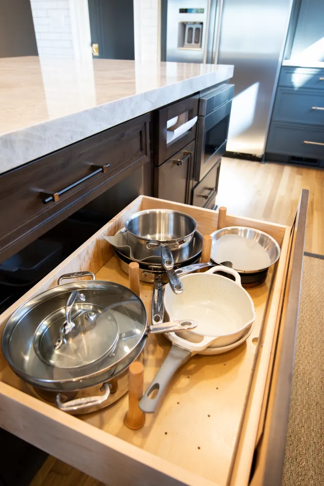 Open kitchen drawer neatly organized with stainless steel pots and pans on wooden pegs under marble countertop.
