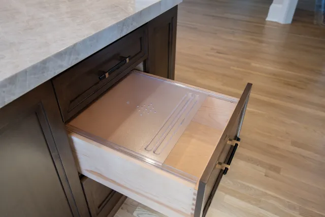 Open wooden kitchen drawer with built-in knife storage and dark cabinetry under marble countertop.