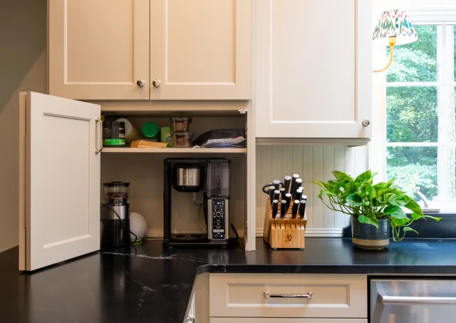 Modern kitchen counter with coffee maker, knife block, green potted plant, and open white cabinet.