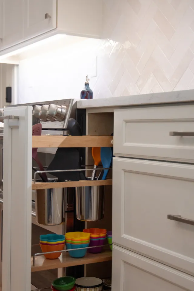 Pull-out kitchen cabinet organizer with colorful bowls, spatulas, and metal containers next to a stove.