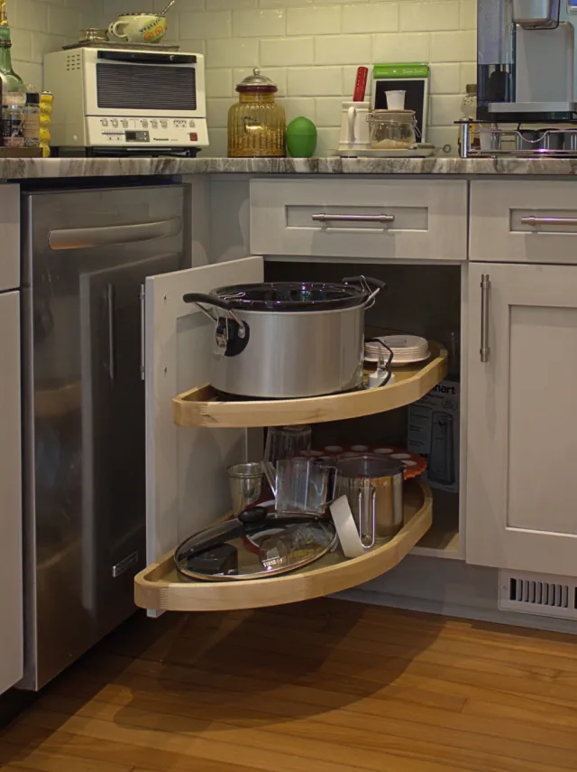 Kitchen corner cabinet with pull-out wooden shelves holding pots, pans, and kitchen appliances on marble countertop.