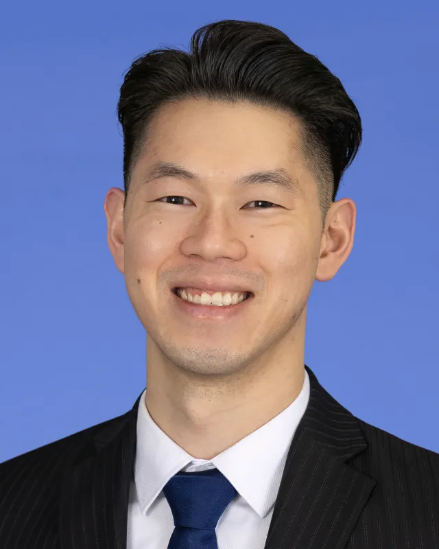 Smiling man in a black pinstripe suit, white shirt, and blue tie against a solid blue background.