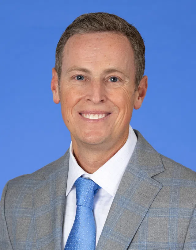 Smiling middle-aged man in gray suit and blue tie against a solid blue background.