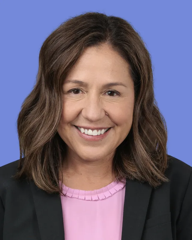 Smiling woman with shoulder-length brown hair wearing a black blazer and pink top against a blue background