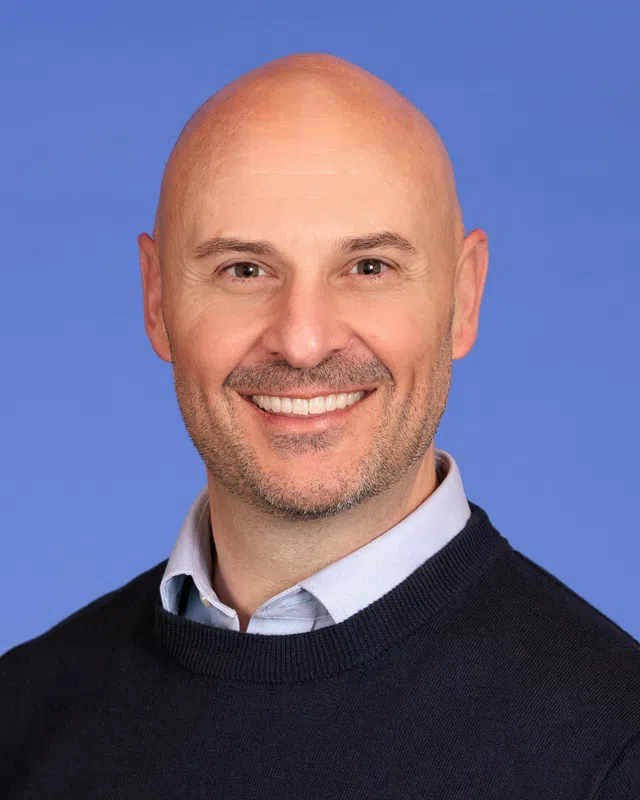Smiling bald man in black sweater and light blue shirt against a solid blue background.