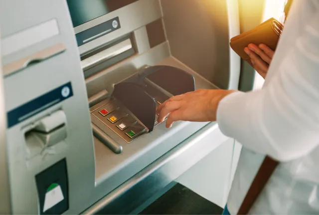 A person using an ATM during daylight, selecting options on the screen with a bank card in hand.