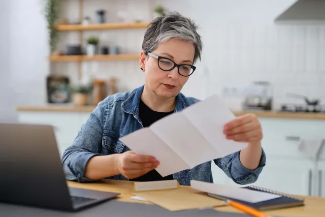 A middle-aged woman with short hair reads documents at a desk with a laptop, showcasing a focused work environment.