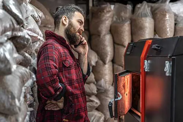 Man in red plaid shirt talking on phone beside pellet stove with sacks of wood pellets in background.