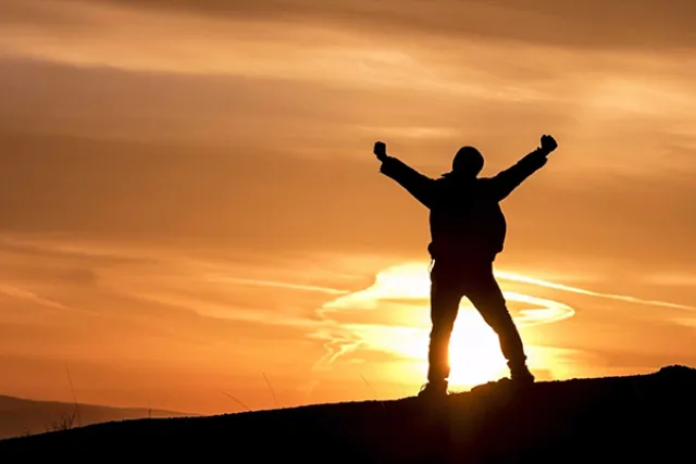 Silhouette of a person with arms raised standing on a hill at sunset with an orange sky.