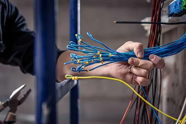 Technician wearing safety gear using a multimeter to repair a wall-mounted air conditioning unit indoors.
