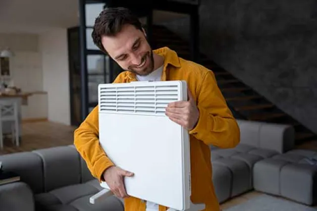 Smiling man in yellow jacket holding a white heater in a modern living room with gray sofas and staircase.