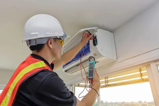 Technician wearing safety gear using a multimeter to repair a wall-mounted air conditioning unit indoors.
