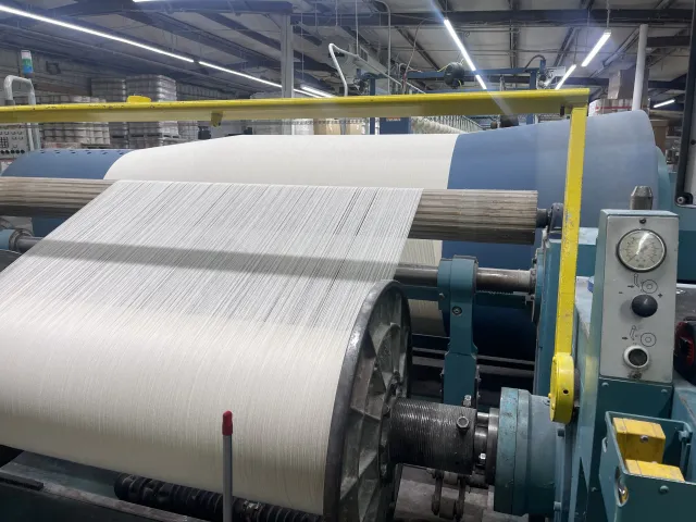 Textile manufacturing machinery winding white fabric yarn in industrial factory setting.