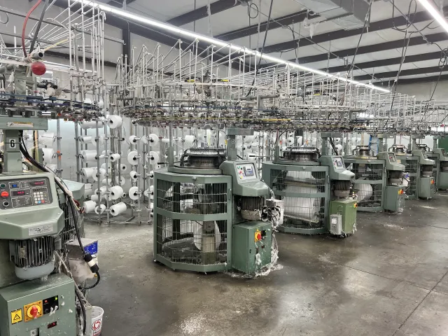 Row of industrial circular knitting machines in a textile factory producing white yarn fabric.