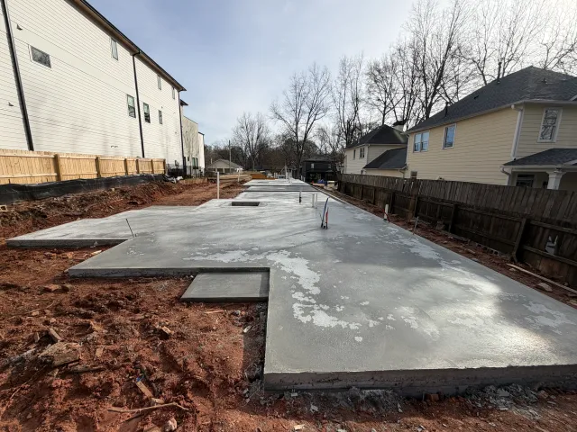 Freshly poured concrete foundation slab for a residential building surrounded by soil and nearby houses.