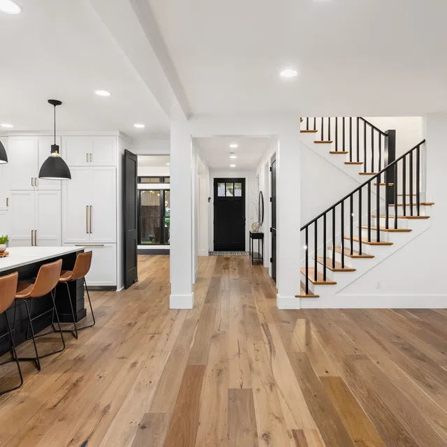 Modern open kitchen and living space with wood floors, black pendant lights, white cabinetry, and a staircase with black railing.
