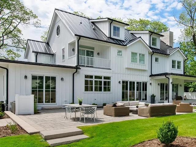 Modern white farmhouse with large patio, outdoor furniture, green lawn, and clear blue sky.