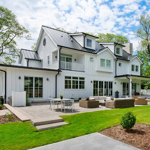 Large modern white farmhouse with metal roof, spacious patio, outdoor furniture, lush green lawn, and surrounding trees.