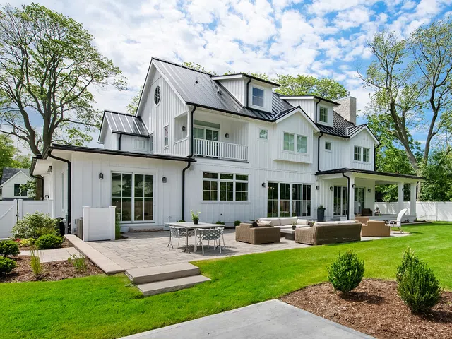 Modern white farmhouse with metal roof, spacious patio with outdoor furniture, and lush green lawn on a sunny day.