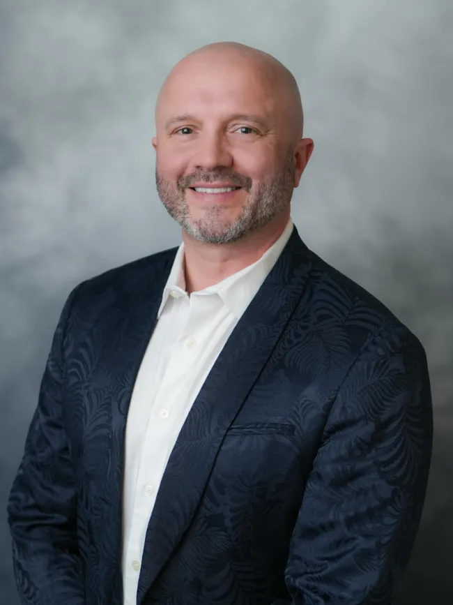 Professional headshot of a smiling man in a patterned navy blazer against a soft gray background.