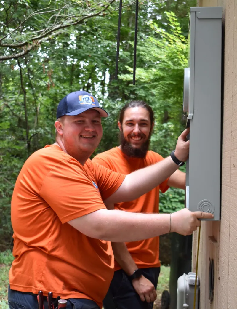 Two Estes Services Technicians workers in orange shirts installing  an electrical panel outdoors surrounded by green trees in Alpharetta, Georgia.