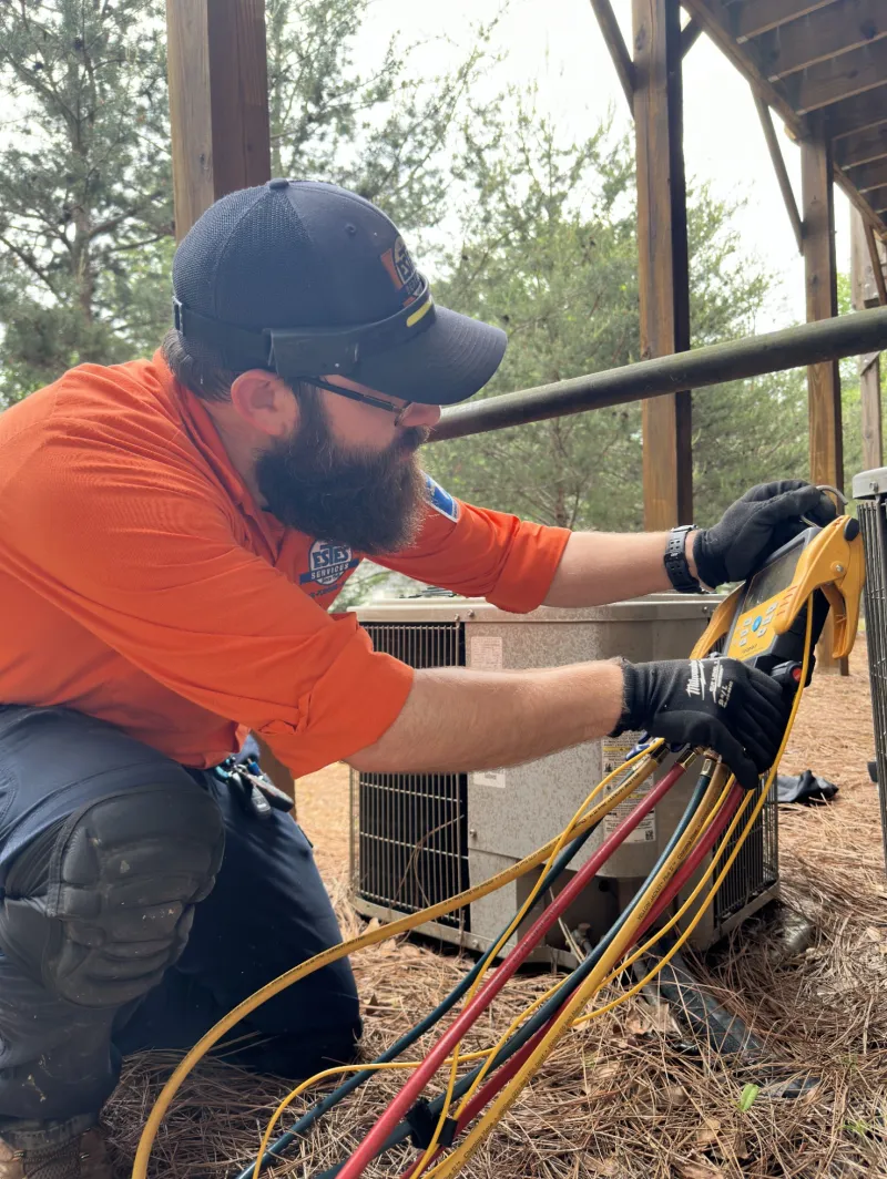Estes Services Technician wearing orange shirt uniform and cap handling hvac gauges electrical cables outdoors near HVAC unit under shelter.