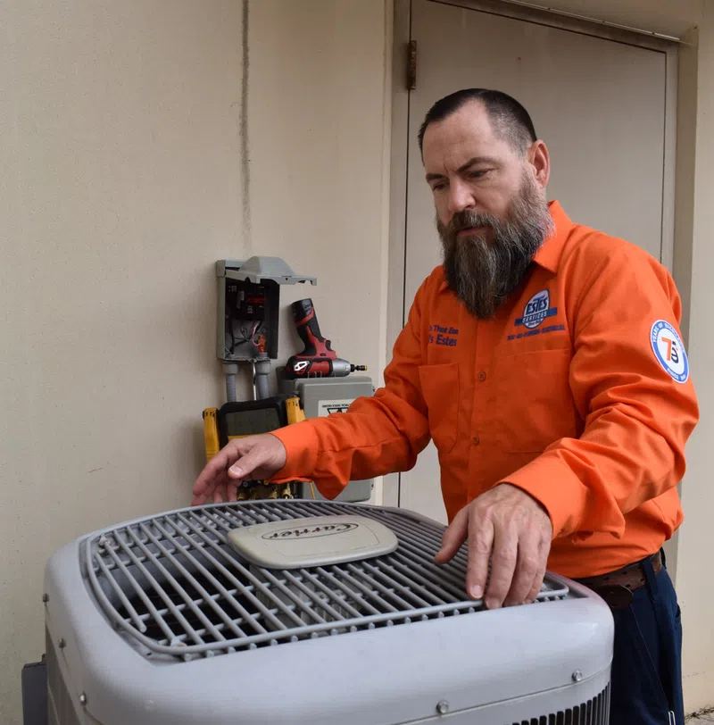 Estes Services Technician in orange uniform inspecting an outdoor air conditioning unit near a beige wall.