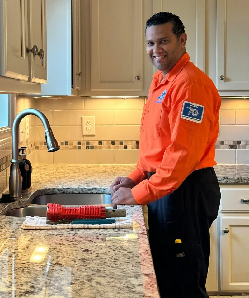 A smiling technician in an orange uniform working at a kitchen sink with tools on the counter.