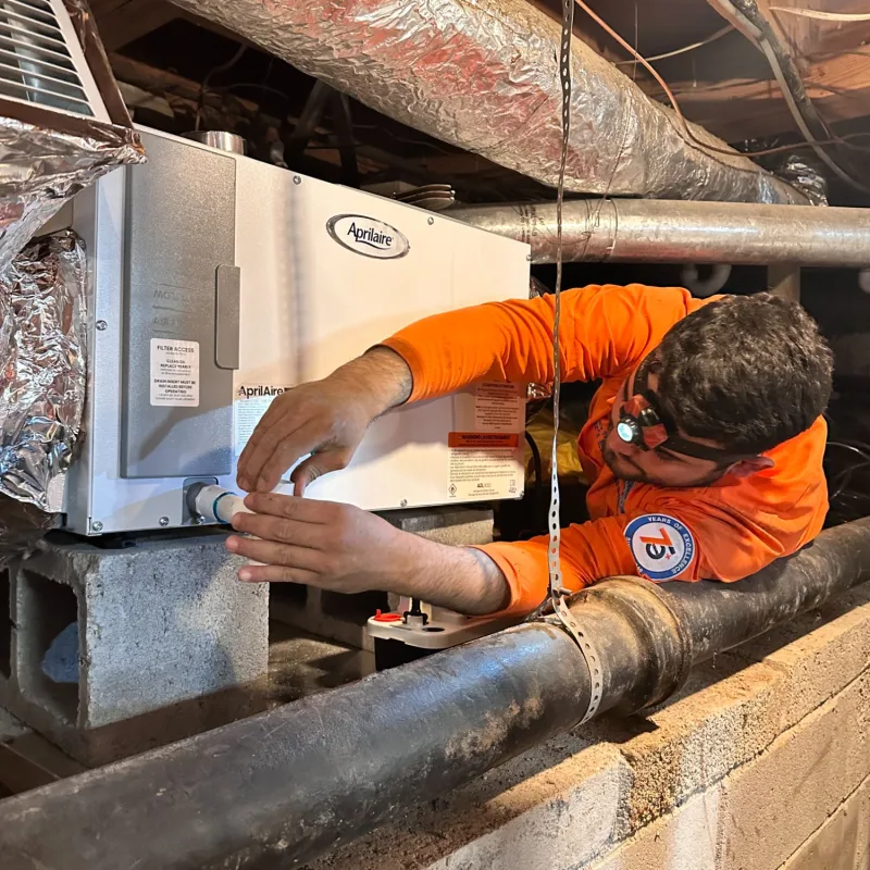 Estes Services Installer in orange shirt uniform installing a Aprilaire Dehumidifier unit in a basement with exposed pipes and ductwork.