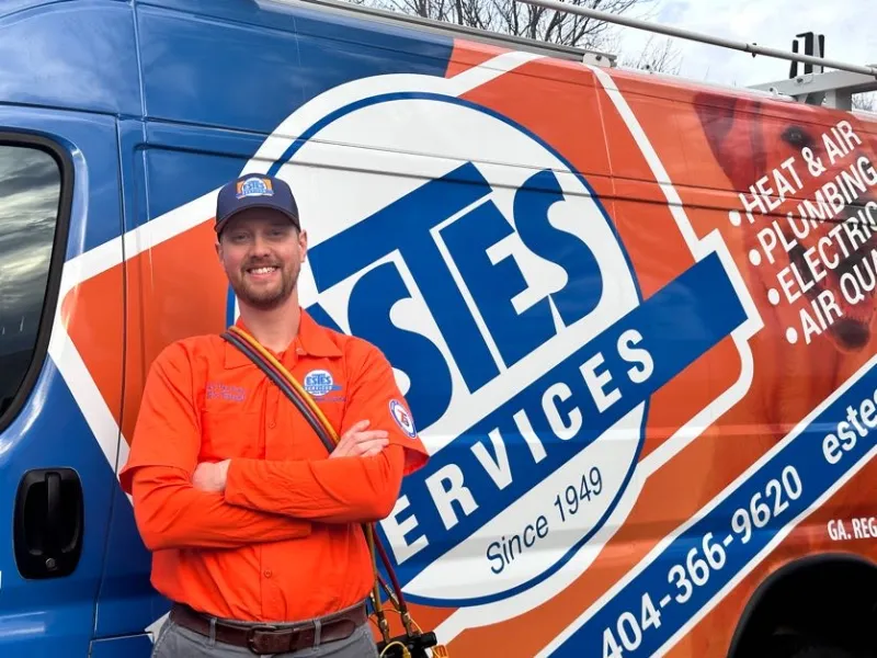 Estes Services HVAC Technician in orange uniform standing with arms crossed in front of Estes Services branded van advertising HVAC and plumbing.
