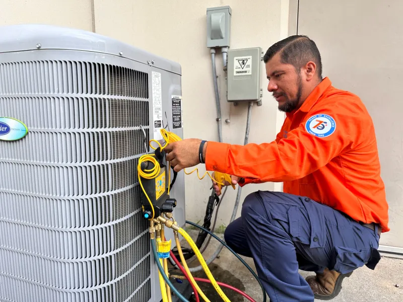 Technician in orange uniform servicing an air conditioning unit using diagnostic tools and gauges outdoors.