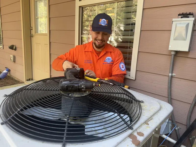 Estes Services Technician in bright orange uniform repairs outdoor air conditioning unit with tools beside a house wall and window.
