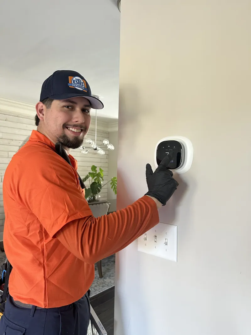 Estes Services Technician in orange shirt and black gloves adjusting a smart thermostat on a beige wall in a modern home.