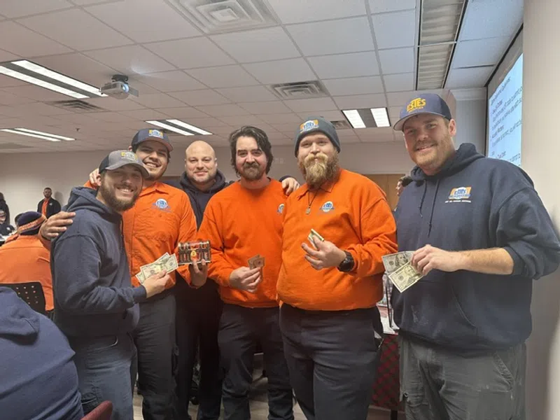 Group of six men in orange and navy shirts holding cash and smiling in a meeting room with projector and chairs.