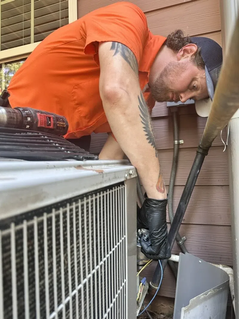 Estes Services Technician in orange shirt repairing outdoor HVAC unit wearing black gloves and using tools by house exterior.