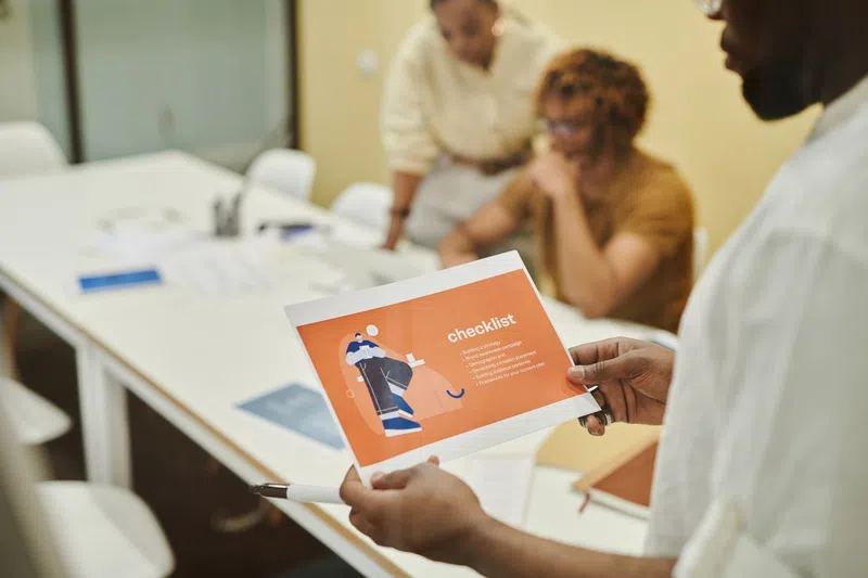 Person holding an orange checklist paper in a meeting with two colleagues at a white table with notebooks.