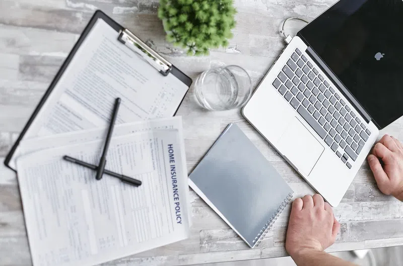 Person working at a desk with a laptop, home insurance policy documents, notebook, and glass of water.