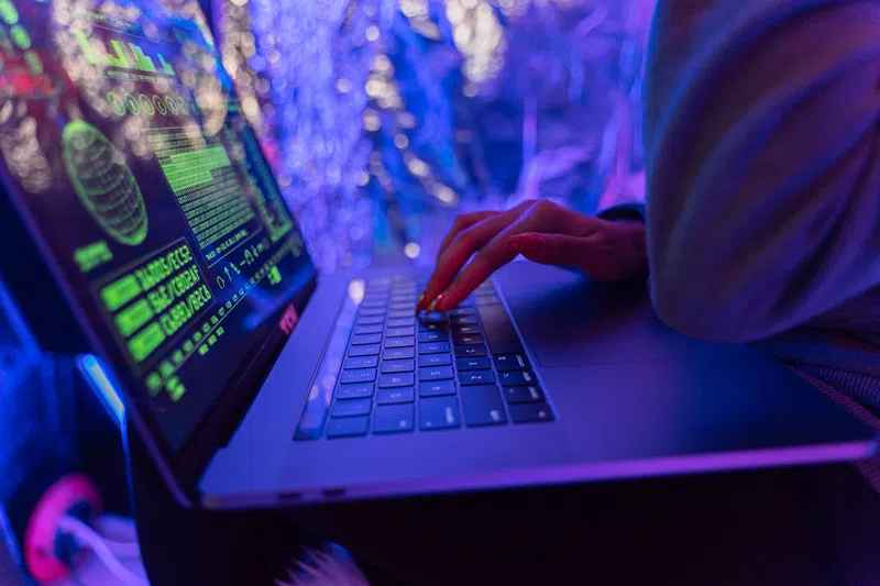 Person typing on a laptop displaying green cybersecurity code in blue and purple ambient lighting.