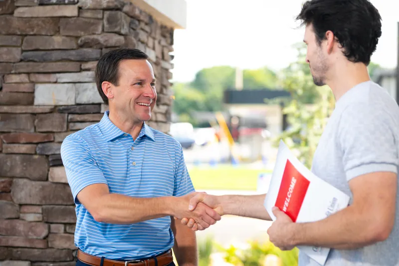 Two men shaking hands outside a building, one holding a welcome brochure, smiling and greeting.