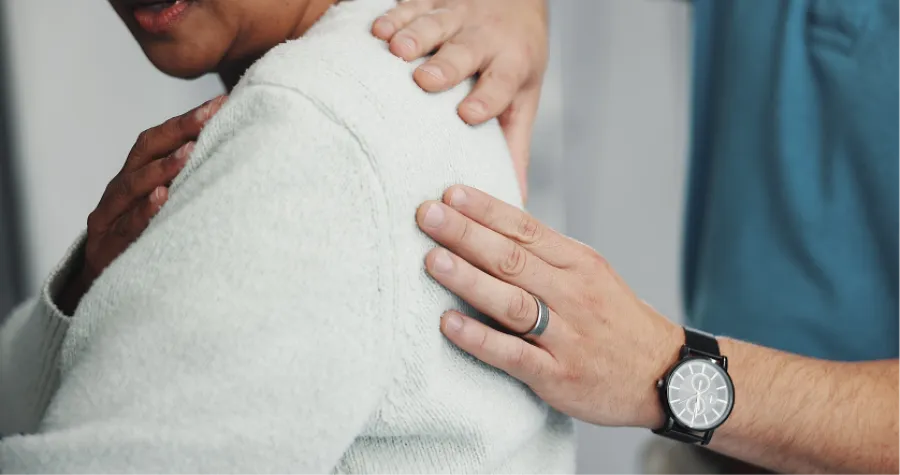 Therapist providing shoulder support and comfort to a patient wearing a white sweater in a clinical setting.