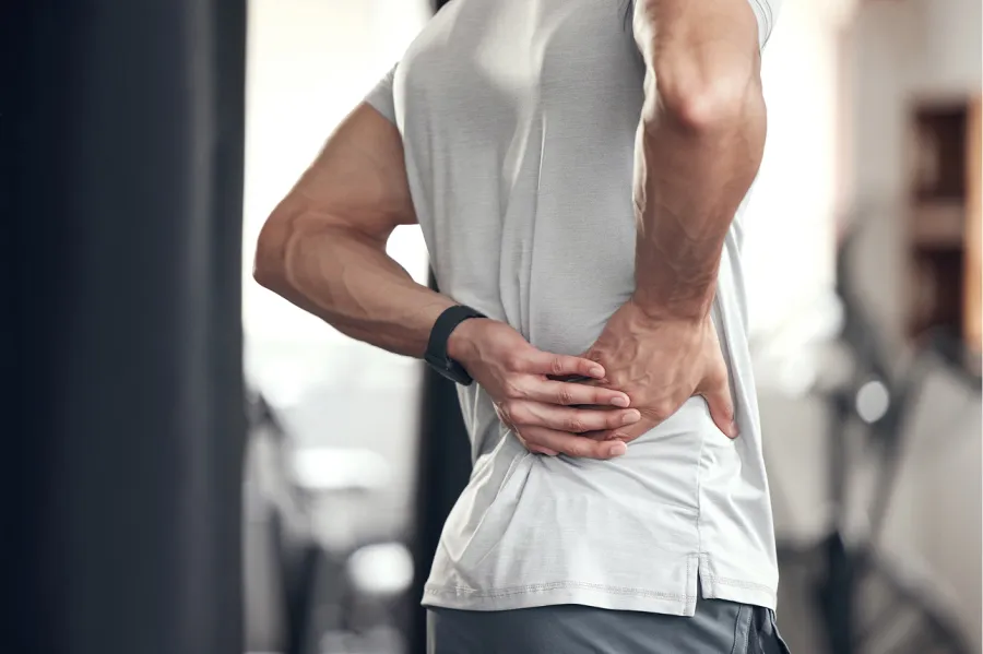 Man wearing white shirt holding lower back in pain, possibly due to injury or strain indoors.