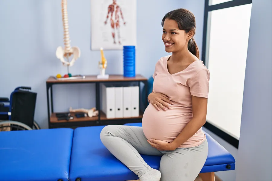 Smiling pregnant woman sitting on medical examination table in clinic, holding belly and looking to side.