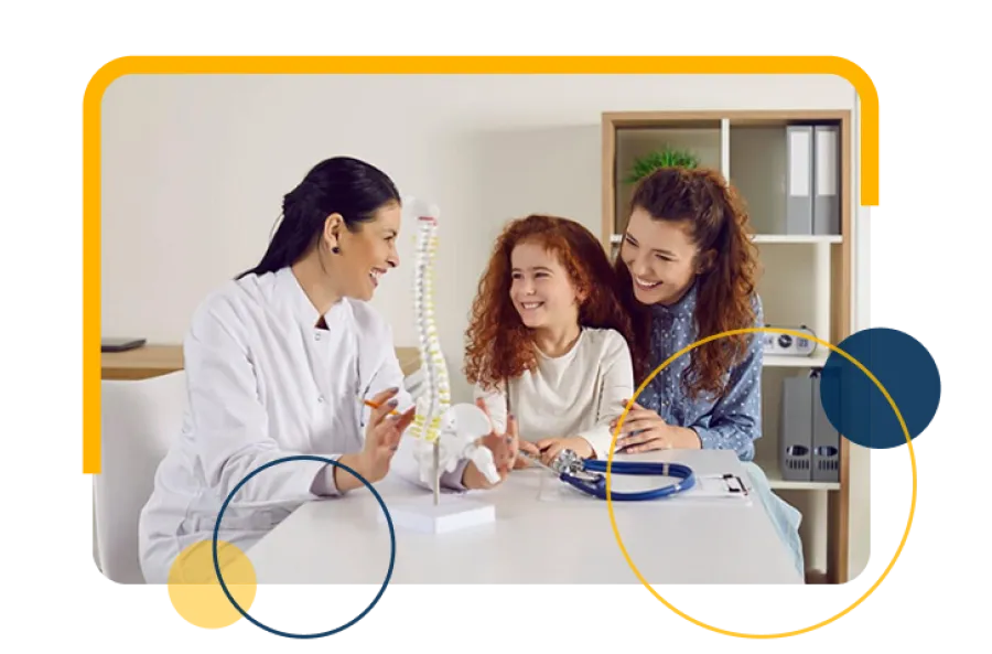 Doctor explaining spine model to smiling girl and her mother in a medical office consultation.